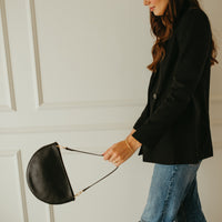 Woman in black blazer and blue jeans holding a black handbag against a white paneled wall.