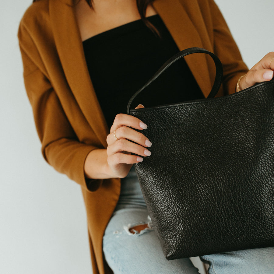 Person holding a black leather handbag wearing a brown coat and light blue jeans.