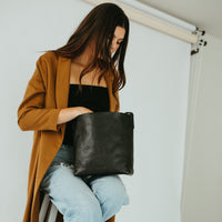 Woman sitting on a stool holding a black leather clutch against a white background