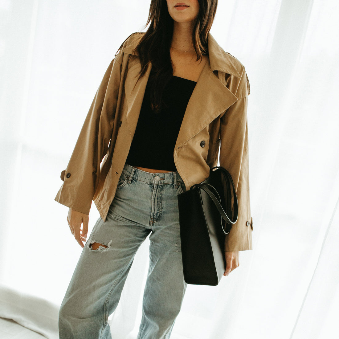 Person wearing a beige trench coat, black top, and light blue jeans on a white background
