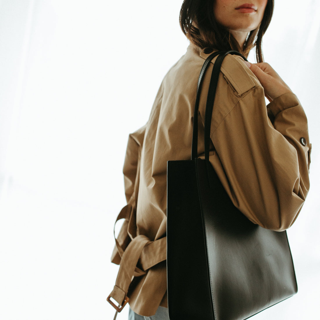 Woman in a beige trench coat holding a black handbag against a white background