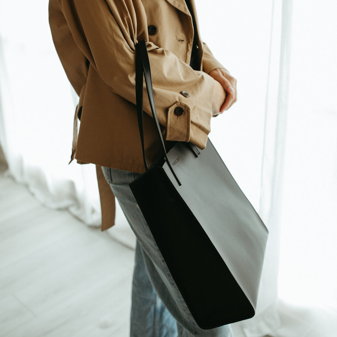 Person wearing a beige trench coat holding a black bag indoors.