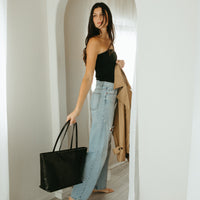 Woman holding a black tote bag and a beige coat, standing in a doorway.
