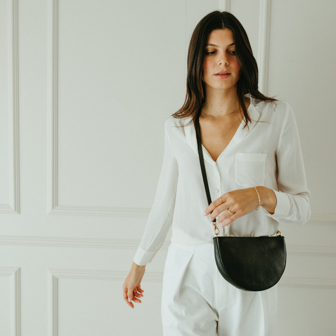 Woman in a white outfit holding a black handbag against a white paneled wall.