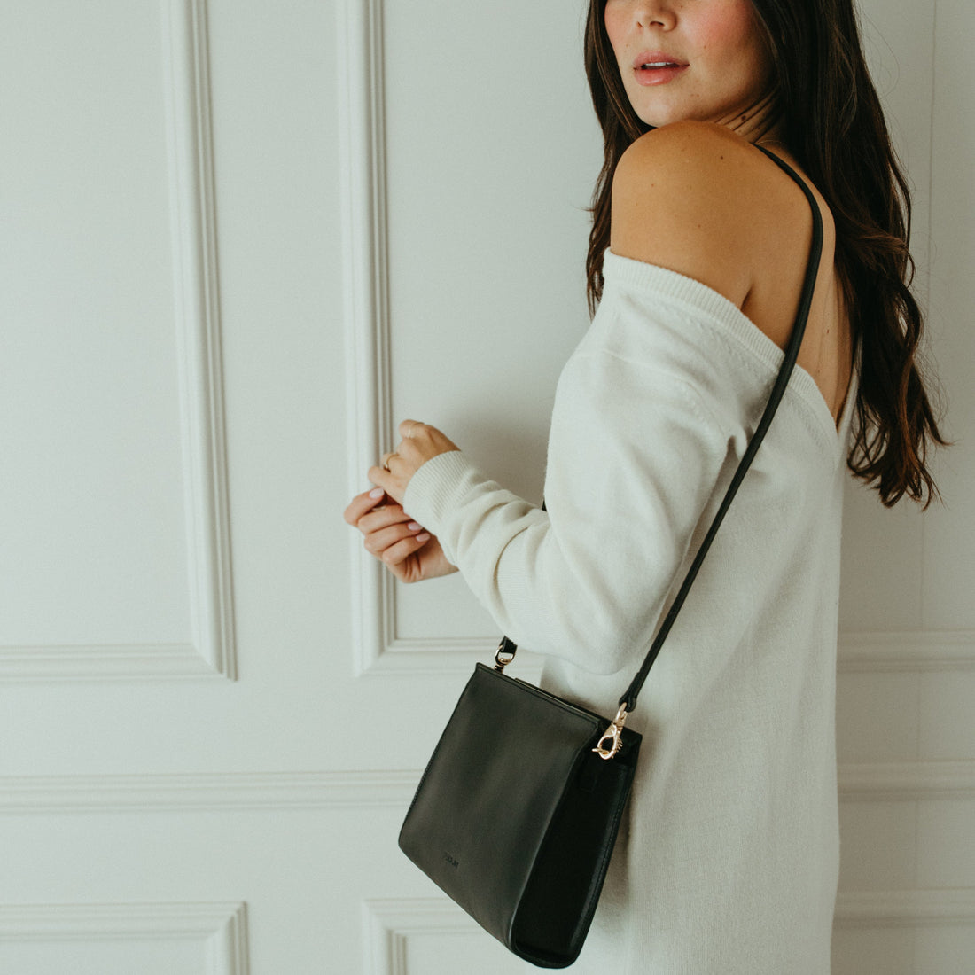 Woman in a white dress holding a black handbag against a white paneled wall.