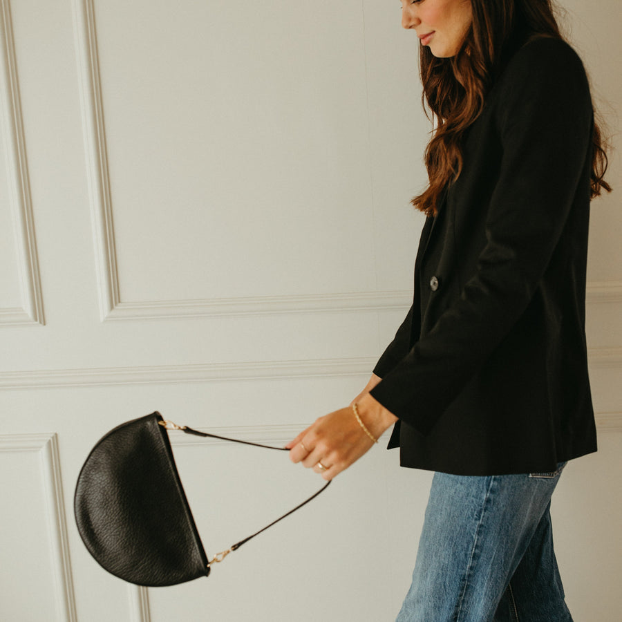 Woman in black blazer and blue jeans holding a black handbag against a white paneled wall.